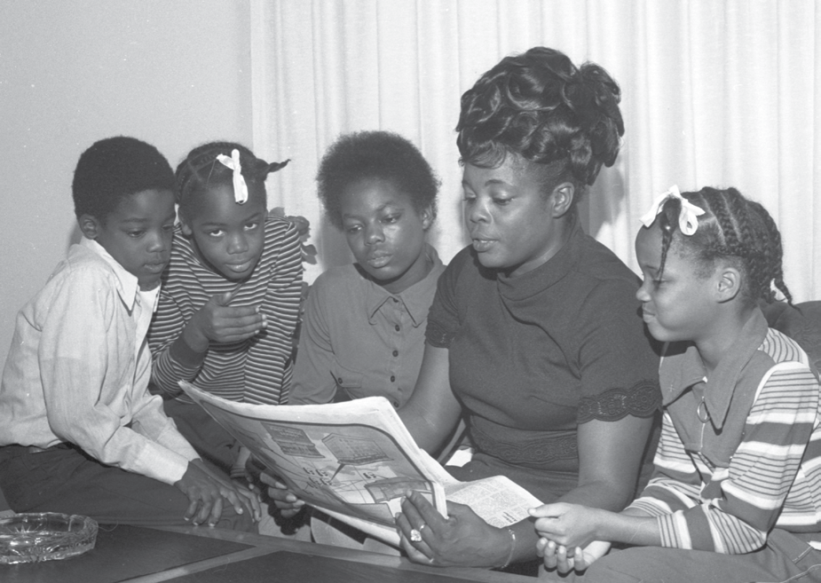 Four children and an adult female read a newspaper together. 